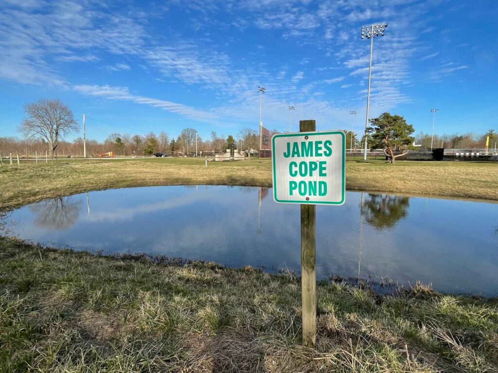 Sign that reads, "James Cope Pond," stationed in front a pond and athletics fields beyond