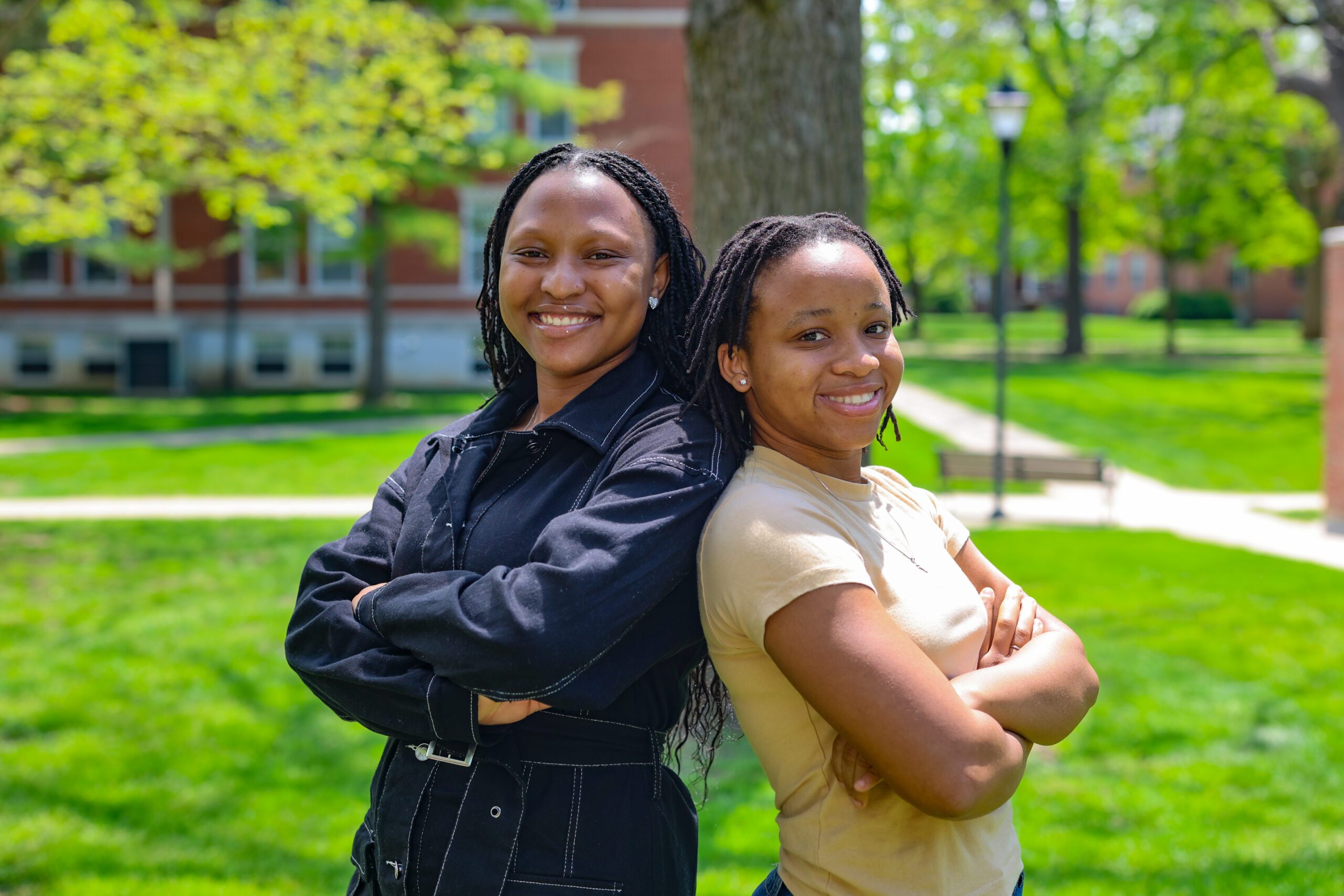 Two young African women stand back to back, arms crossed, smiling at the camera. The woman on the left is slightly taller than the one on the right, and wears a black outfit. The woman on the right wears a pale yellow t-shirt.