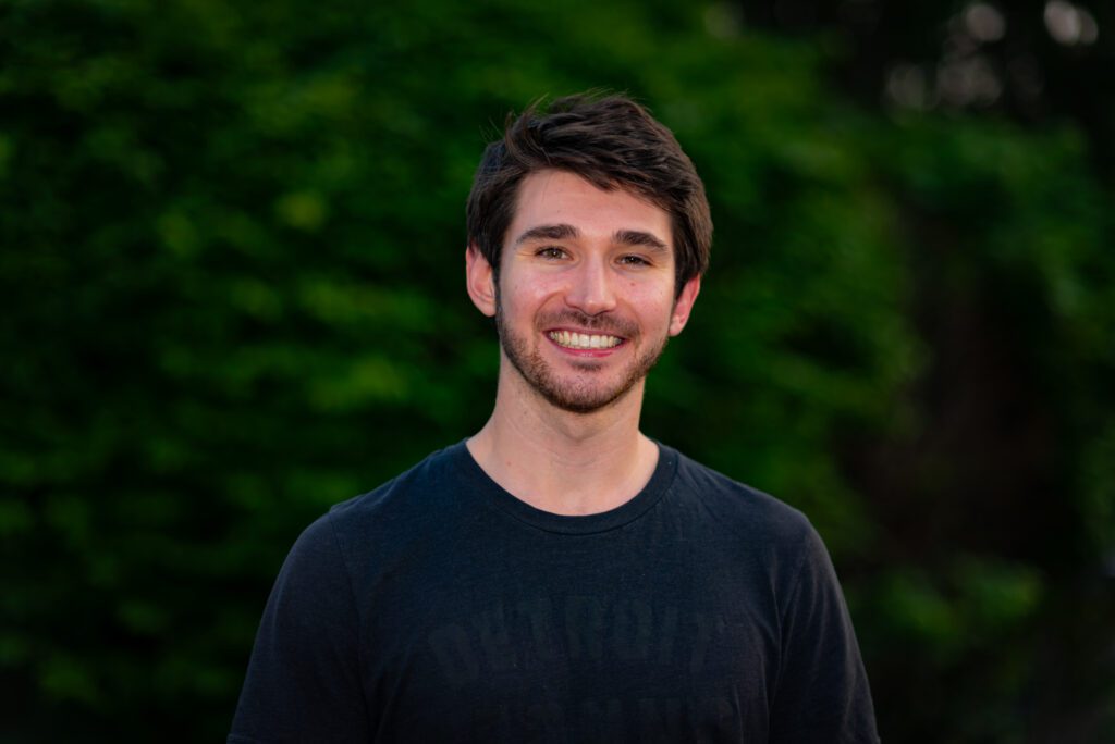Sam Pigott smiles with a green, tree-lined background behind him