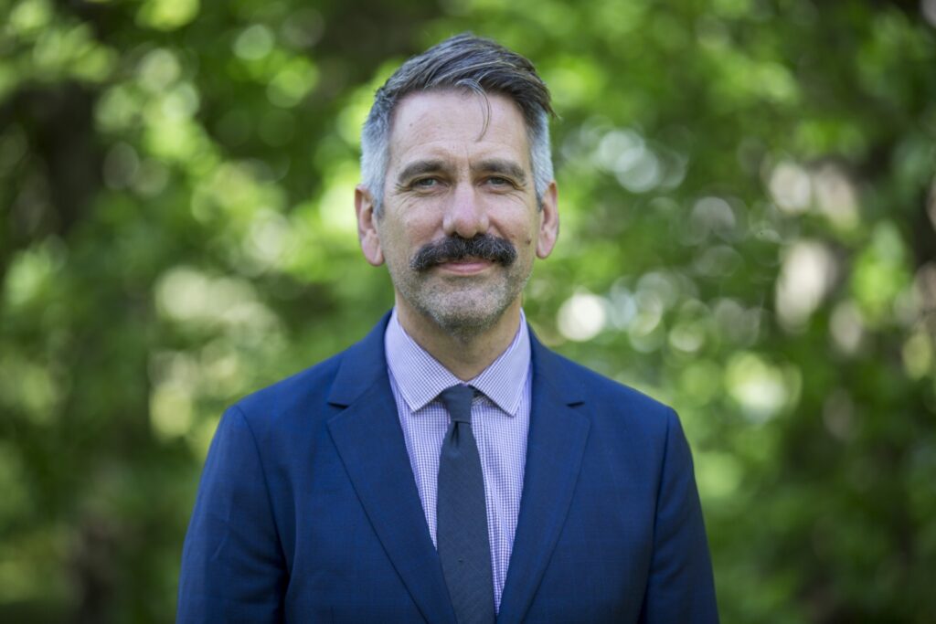 Jeffrey Perkins smiles in front of a blurry backdrop lined by green leafs from a tree. He is wearing a blue sports jacket and tie.