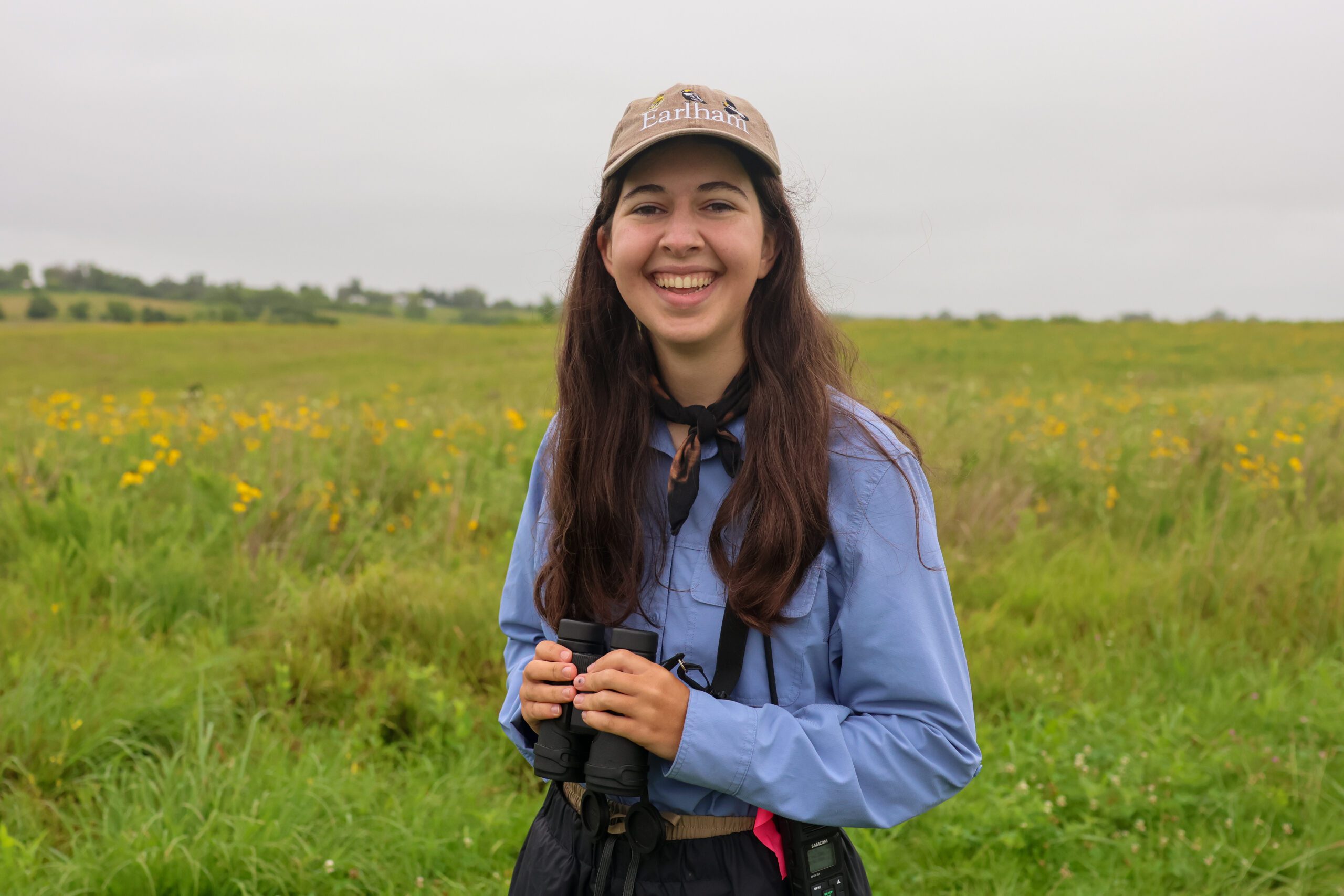 Earlham College student Sarah Osburn smiles while holding binoculars and wearing a hat with the words Earlham and illustrations of birds on it.