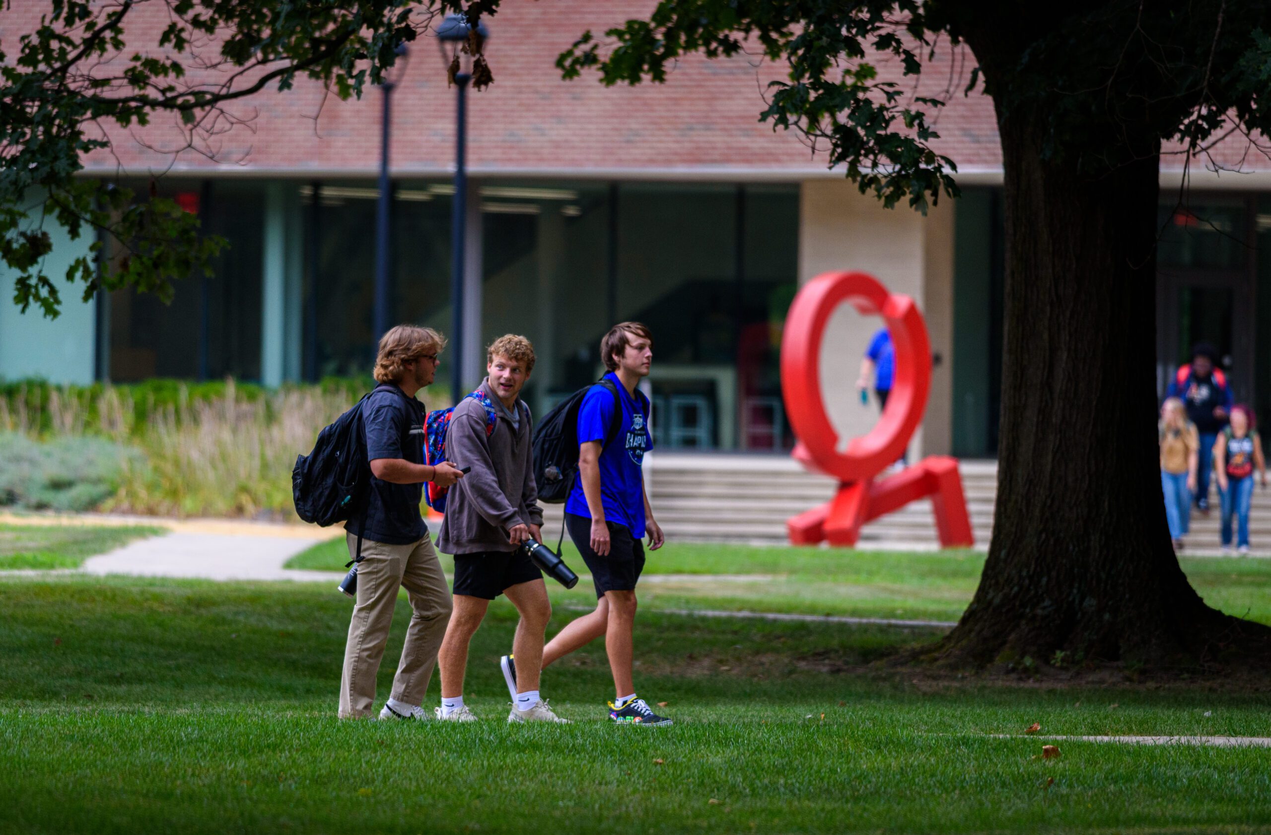 Three male students walk across Earlham College's campus with a red sculpture in the shape of an interlocking X and O in the background.