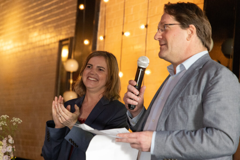 Olivia Ramsey is pictured on the left side of the image clapping while wearing a dress while Fabrice Jaumont holds a microphone to her right.