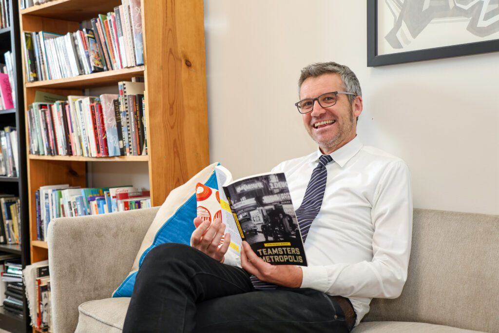 Ryan Murphy sitting on a couch in his office with his legs crossed while clutching a copy of his new book.