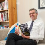 Ryan Murphy sitting on a couch in his office with his legs crossed while clutching a copy of his new book.