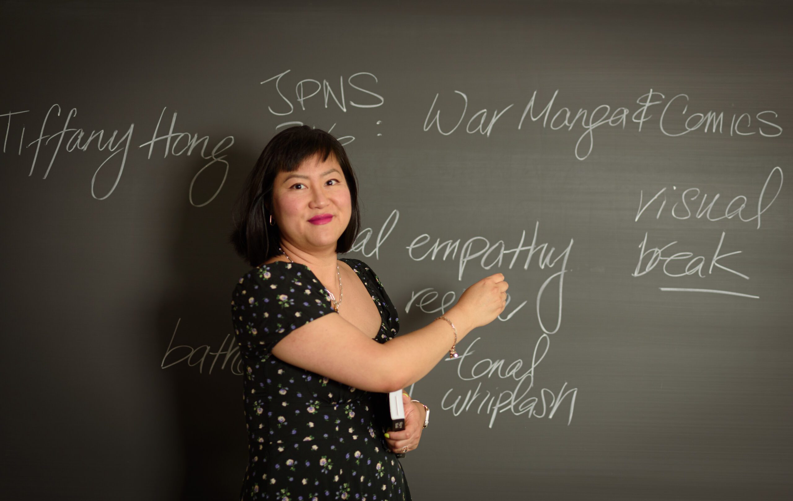 Earlham College professor Tiffany Hong smiles in front of a chalkboard with a piece of chalk in her hands
