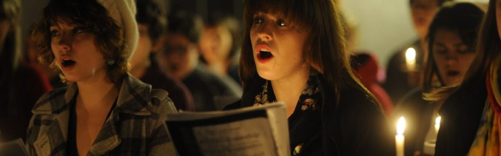 A young woman with brown hair and bangs sings with her mouth open, holding a lit candle. Another woman is partially visible to her left, also singing. People in the background are out of focus. The scene is lit by candlelight.