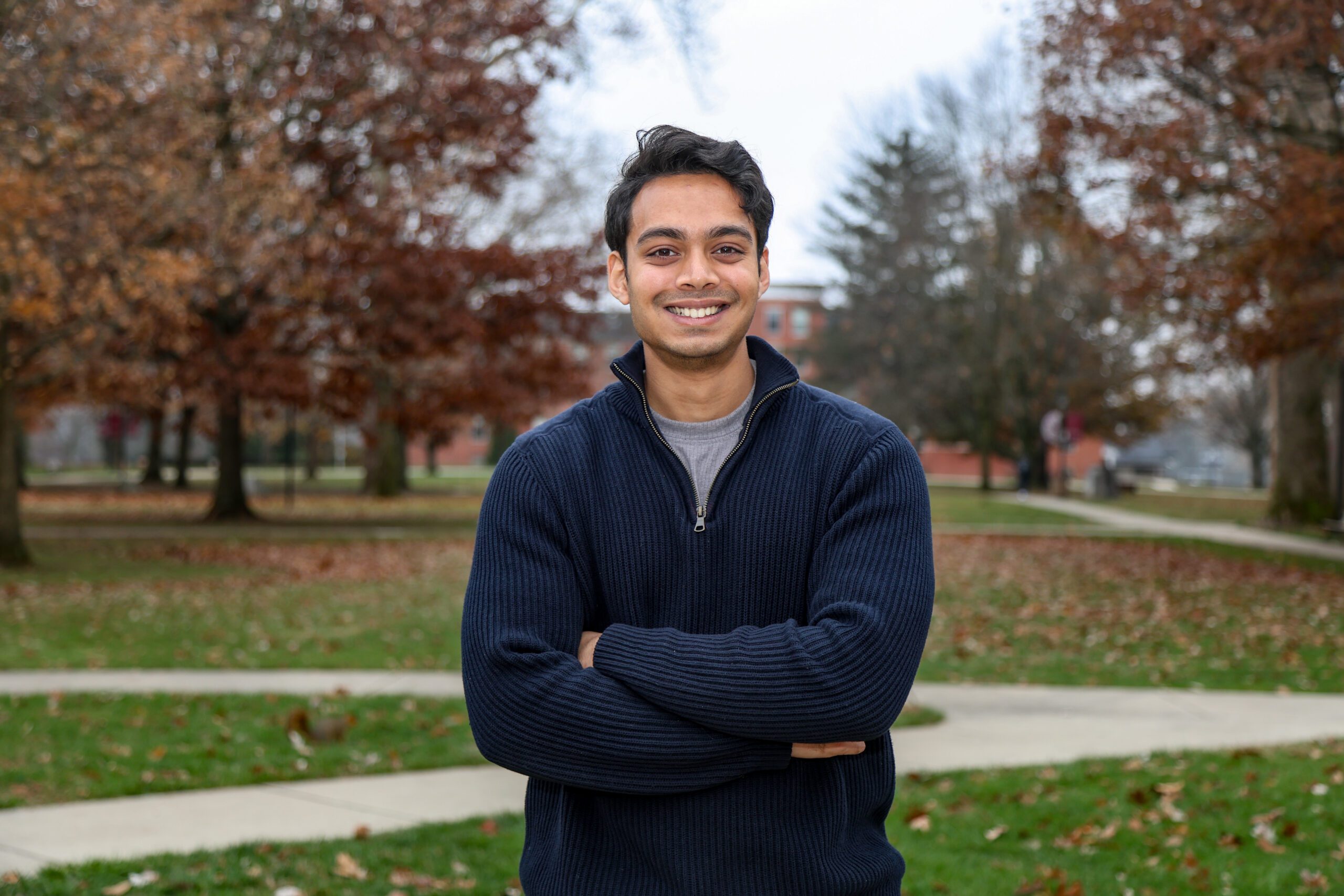 Rayyan Afzal stands with his arms crossed wearing a blue coat with a fall landscape on Earlham College's campus behind him.