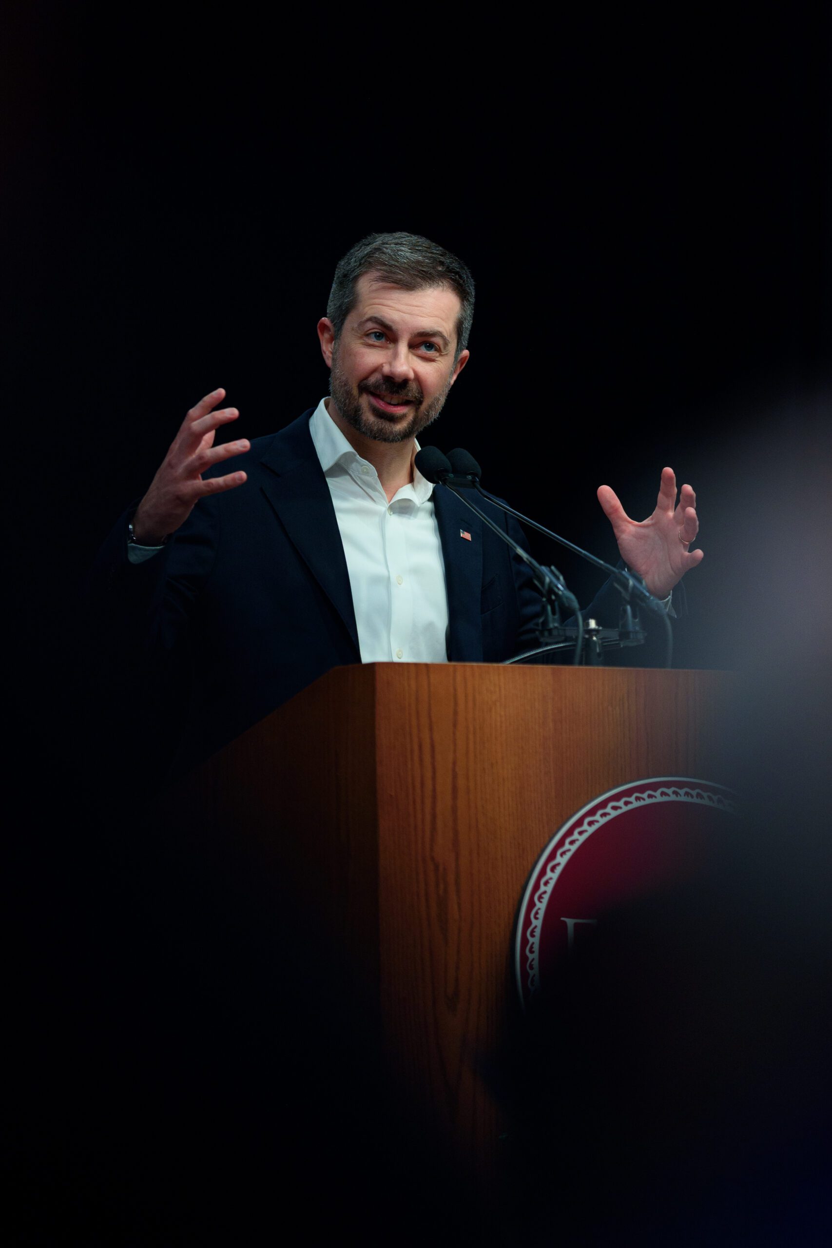 Pete Buttigieg speaks behind a podium with both of his hands outstretched