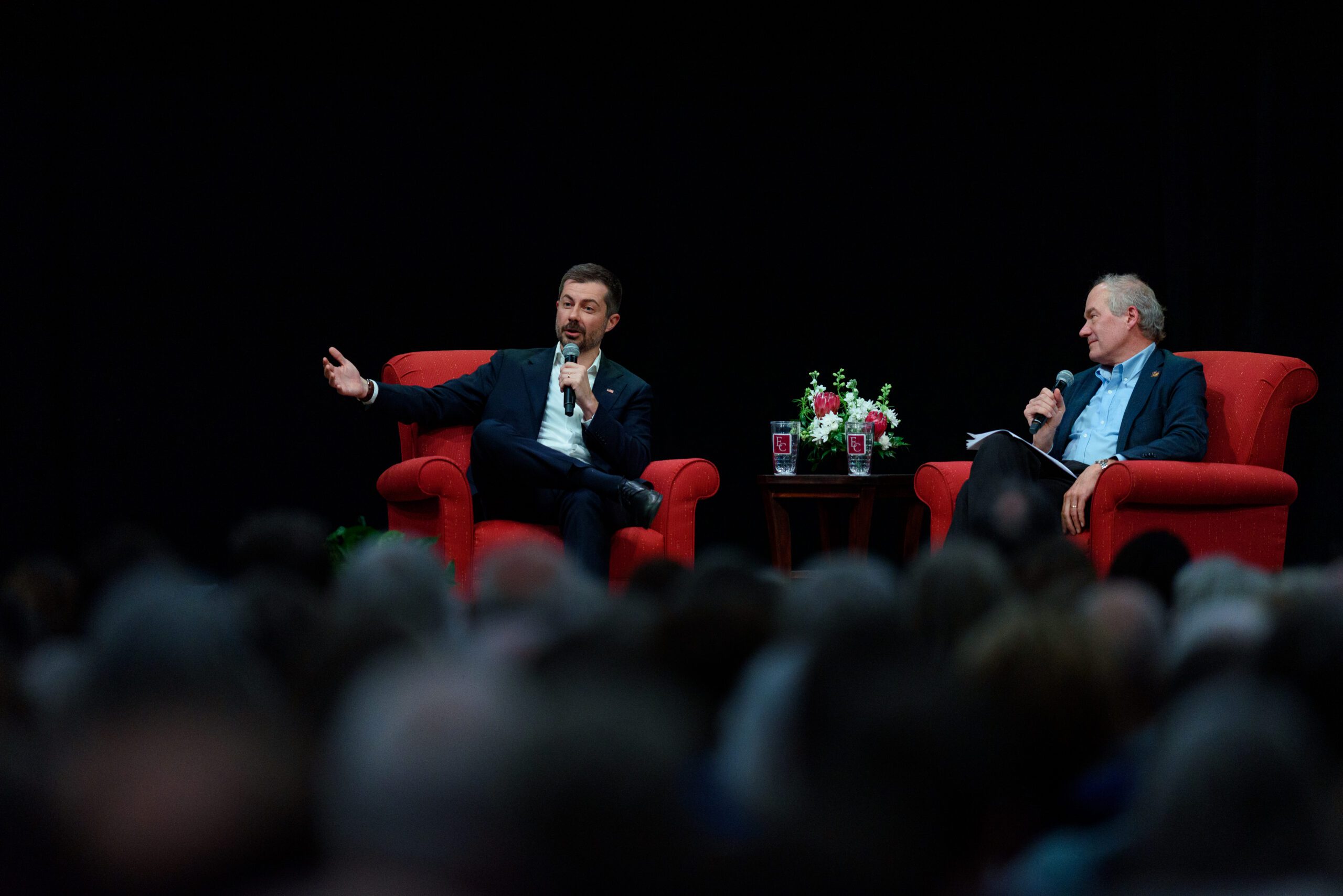 Pete Buttigieg and Paul Sniegowski seated in red chairs with the backsides of audience members blurred in foreground.