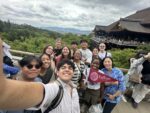 A group of Earlham College students pose for a photo outside in Japan with a student in the middle holding an Earlham pennant.