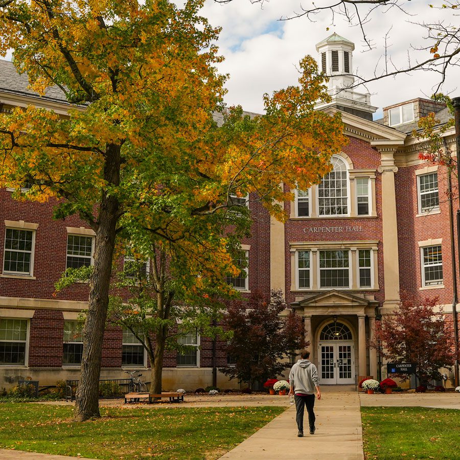 Three male students walk across Earlham College's campus with a red sculpture in the shape of an interlocking X and O in the background.