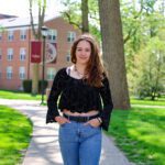 Briana Ancalmo stands on a paved sidewalk on the Earlham College campus with her hands in her pocket