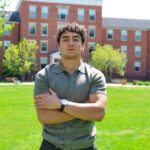 Armen Sargsyan stands in front of Earlham Hall wearing a gray polo shirt and with his arms crossed