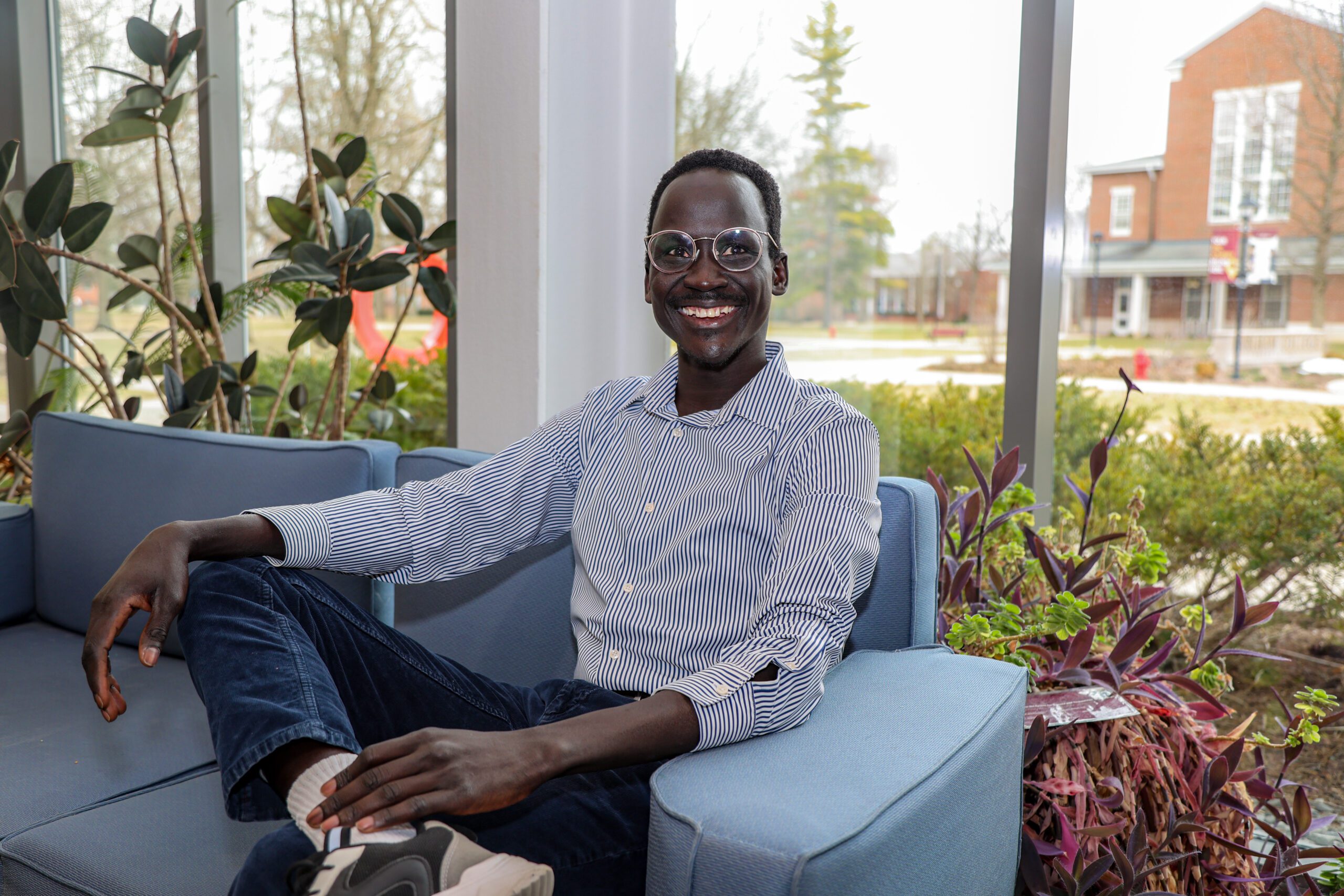 Malou Cithiec sits in a blue chair with his legs crossed surrounded by plants