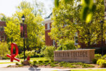 Green leaves from trees, lightpole banners, and a red metallic sculpture surround a stone sign with the words Earlham College in it
