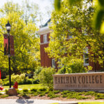 Green leaves from trees, lightpole banners, and a red metallic sculpture surround a stone sign with the words Earlham College in it
