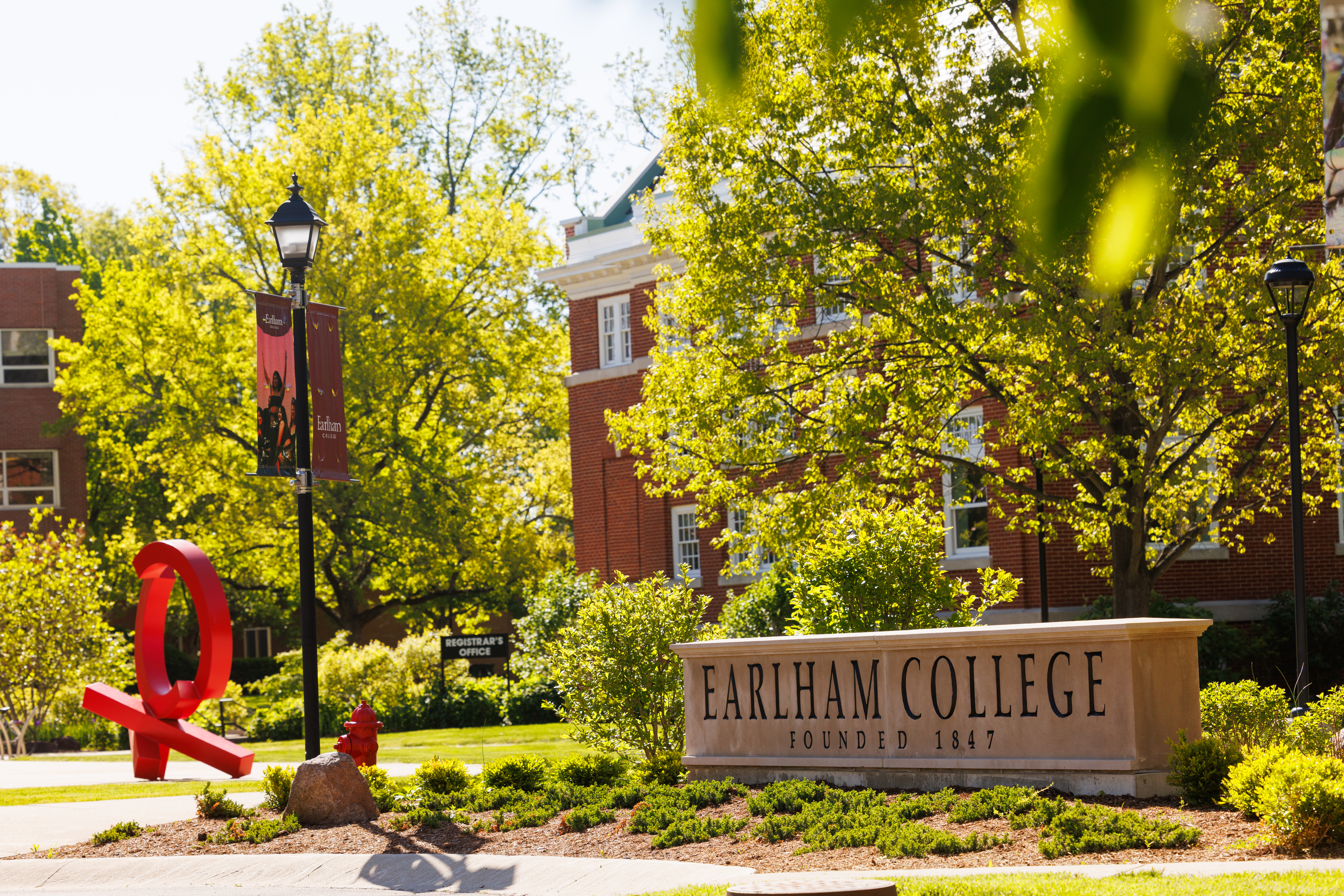 Green leaves from trees, lightpole banners, and a red metallic sculpture surround a stone sign with the words Earlham College in it