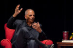 Eric Holder sits on a red couch gesturing with his hands and look toward the right