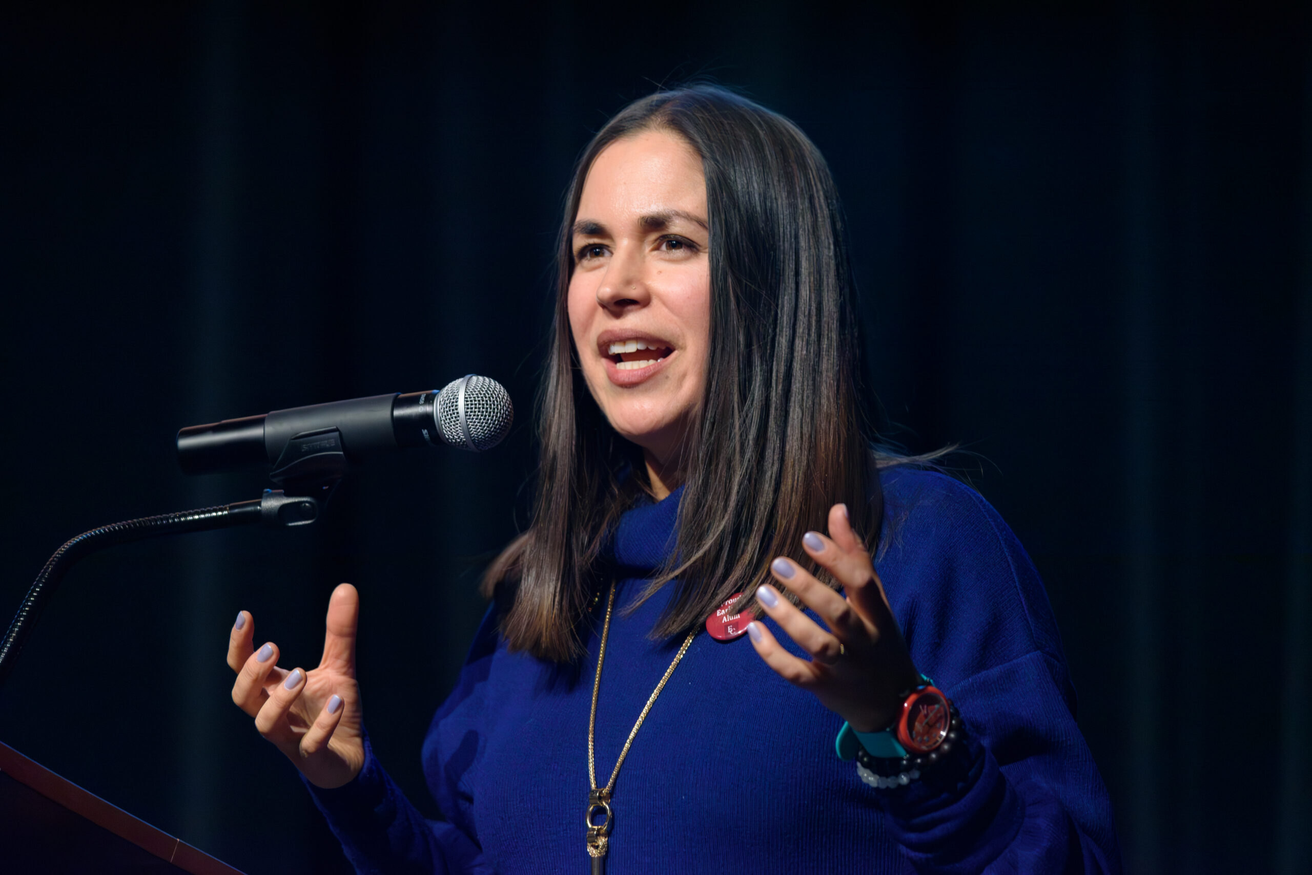 Katie Yamasaki stands behind a microphone with both of her hands gesturing.